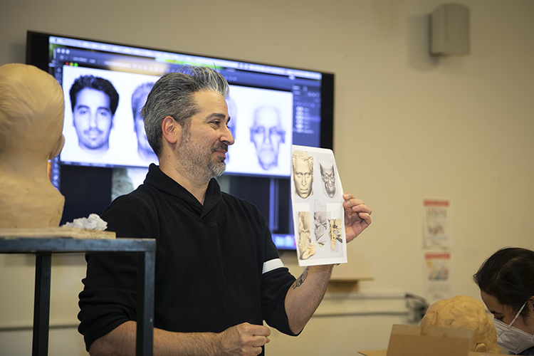 Joe Mullins stands in front of the sculpture class facing the students with a screen behind him showing facial reconstruction techniques